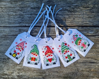 May include: Five white gift tags with cross-stitched gnome designs. Each tag features a different gnome wearing a hat and holding a gift or lantern. The gnomes have white beards and are set against a white background. White ribbons are attached.