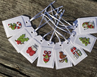 May include: A collection of white gift tags with various Christmas-themed cross-stitch designs. The tags feature images of a candy cane, holly, a snowman, a Christmas tree, gifts, and other festive elements, all with white ribbon.