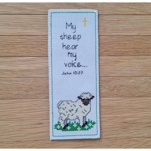 Pode incluir: Um marcador de livro feito à mão em ponto cruz com o texto "As minhas ovelhas ouvem a minha voz... João 10:27". O marcador de livro apresenta uma ovelha branca com rosto e pernas pretas, em pé sobre relva verde. A borda é verde.