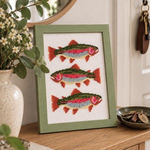 May include: A framed cross-stitch embroidery featuring three trout fish arranged vertically. Each fish is stitched with green, pink, and light blue thread on a white fabric background. The frame is a simple, light green wood with a textured finish.
