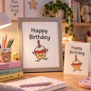 Può includere: Un ricamo a punto croce incorniciato e un biglietto d'auguri abbinato, entrambi con il testo Happy Birthday e un'illustrazione colorata di un cupcake con una candela accesa. Il cupcake ha una base gialla, glassa rossa, braccia e gambe.