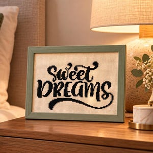 May include: A rectangular cross-stitch artwork in a light green frame featuring the black text Sweet Dreams on a cream-coloured fabric background. The embroidery displays a decorative script font with a flourish under the word Dreams.