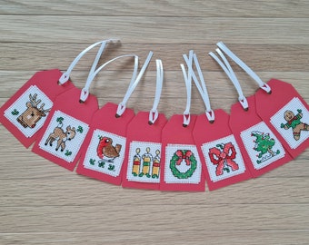 May include: A collection of eight red gift tags with white ribbon, each featuring a cross-stitch design. The designs include a reindeer, deer, robin, candles, wreath, bow, Christmas tree, and gingerbread man. Perfect for holiday gifting.
