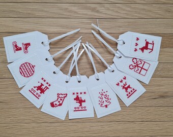 May include: Ten white gift tags with red cross-stitch designs of various Christmas icons, including a stocking, a tree, a reindeer, a present, and a bell. The tags are arranged in a circle on a wooden surface.