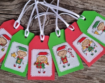 May include: Six Christmas gift tags with cross-stitch designs of elves wearing Santa hats. The tags are red and green with white ribbon.