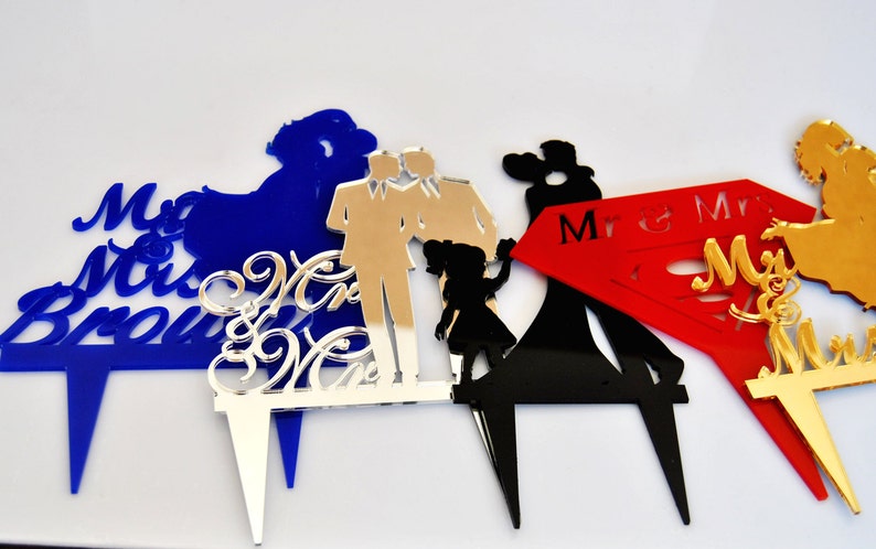 May include: Four different cake toppers made of acrylic. The first topper is blue with the text "Mr & Mrs Brown". The second topper is silver with the text "Mr & Mrs". The third topper is black with a couple dancing and the text "Mr & Mrs". The fourth topper is gold with a couple dancing and the text "Mr & Mrs".