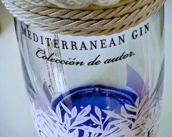 May include: A clear glass bottle of Gin Mare with a blue base and white botanical design. The bottle is labeled "MEDITERRANEAN GIN" and "Colección de autor." A rope detail adorns the top. The bottle contains 700 ml and is 42.7% alcohol.