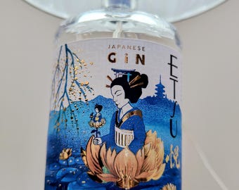 May include: A clear glass bottle of Etsu Japanese Gin with a blue and gold label. The label features an illustration of a person in a blue kimono sitting in a large gold lotus flower, surrounded by blue water lilies, a torii gate, and a pagoda.