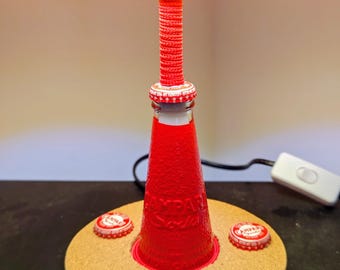 May include: A table lamp featuring a red Campari Soda bottle as the central base, mounted on a circular cork stand decorated with three Campari Soda bottle caps. The lamp has a red textured stem and a yellow lampshade with a white power cord.