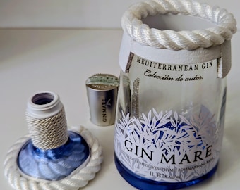 May include: A clear glass bottle of Gin Mare Mediterranean Gin with a blue base and white rope detailing. The label features white botanical illustrations and text. The bottle is partially open, with the cap and a small metal top beside it.