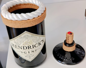 May include: A black gin bottle with a white label and a cork stopper. The bottle has a white rope detail around the neck. The stopper has a cork base and a black top with the brand name. The bottle is 1 litre.
