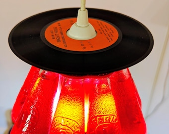 May include: A distinctive pendant light, crafted from recycled materials. The lamp features a black vinyl record as the top shade, and a cluster of red glass bottles forming the lower shade. The bottles are lit, producing a warm, red glow.