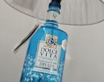 May include: A table lamp constructed from a bright blue glass Dolce Vita Dry Gin bottle. The lamp features a white pleated fabric lampshade, a black socket, and a white power cord with an inline switch. The bottle label displays text including Dolce Vita, Mediterranean Distilled Dry Gin, and Tribute to Capri.