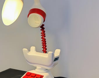 May include: A white and red telephone-shaped desk lamp. The lamp has a red coiled cord, a red keypad, and a red and white lamp head. The light is on, illuminating the area. The lamp is on a black surface.
