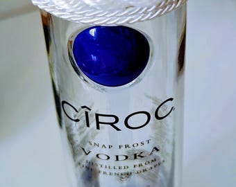 May include: A clear glass Cîroc vodka bottle with a white rope detail around the top. The bottle has a blue liquid at the bottom and a blue circular design. The words "Cîroc" and "Vodka" are visible on the bottle.