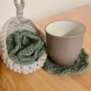 May include: Two crocheted coasters in a sage green color, one with a handle, and a gray ceramic mug sitting on top of the coaster.