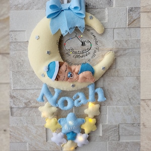 Può includere: Una luna crescente gialla con un bambino che dorme dentro, un nome in feltro blu "Noah" con luci scintillanti e una ghirlanda di stelle gialle con luci appese sotto. La luna ha il testo "fatto a mano con amore" e "Boutique by Monika" su di essa.