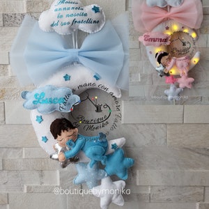 Può includere: Un mobile per neonati in feltro blu e bianco con una luna crescente, stelle e una figura di un bambino che dorme. Il mobile è decorato con il testo "Fatto a mano con amore fatto con amore Boutique by Monika" e "Luca".