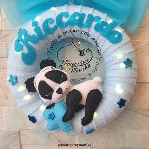 Può includere: Una ghirlanda di feltro blu con il nome "Riccardo" scritto in lettere di feltro blu. La ghirlanda è decorata con un orso panda addormentato che tiene una stella blu e piccole stelle blu con centri bianchi. La ghirlanda è illuminata da luci LED bianche. Il testo "Fatto a mano con amore" è scritto sulla ghirlanda, insieme al testo "Boutique by Monika".