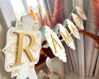 May include: A decorative banner with white and gold accents. The banner features individual lettered panels spelling out a name, with a large gold letter "R" at the beginning. The panels are connected with clear beads. The banner is displayed in front of a window.