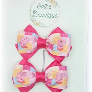 May include: Two pink hair bows featuring a Peppa Pig design. The bows are made from pink and yellow checked ribbon with a Peppa Pig character on each square. The bows are attached to a white card with the text "Salt's Boutique" in a green circle.
