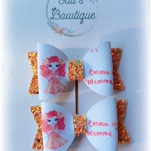 May include: Two white hair bows with orange glitter trim. Each bow features a cartoon girl with red hair and a pink dress. The text "Emma Mamma" is printed on each bow.
