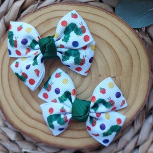 May include: Two green and white hair bows with a green caterpillar and colourful fruit print. The bows are tied with a green ribbon.
