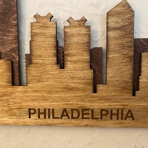 Puede incluir: Arte de pared de madera con la silueta de Filadelfia. La obra de arte est&aacute; hecha de madera en capas con un acabado natural, mostrando los edificios ic&oacute;nicos de la ciudad. La palabra "PHILADELPHIA" est&aacute; grabada en la parte inferior. Una pieza decorativa para el hogar o la oficina.