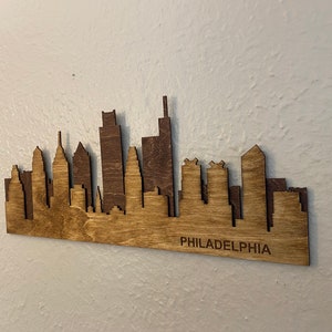 Puede incluir: Pieza de arte de madera con la silueta de Filadelfia. La silueta est&aacute; cortada con l&aacute;ser de madera, con un dise&ntilde;o de dos tonos, utilizando un marr&oacute;n m&aacute;s oscuro para algunos edificios. La palabra "PHILADELPHIA" est&aacute; grabada en la parte inferior.