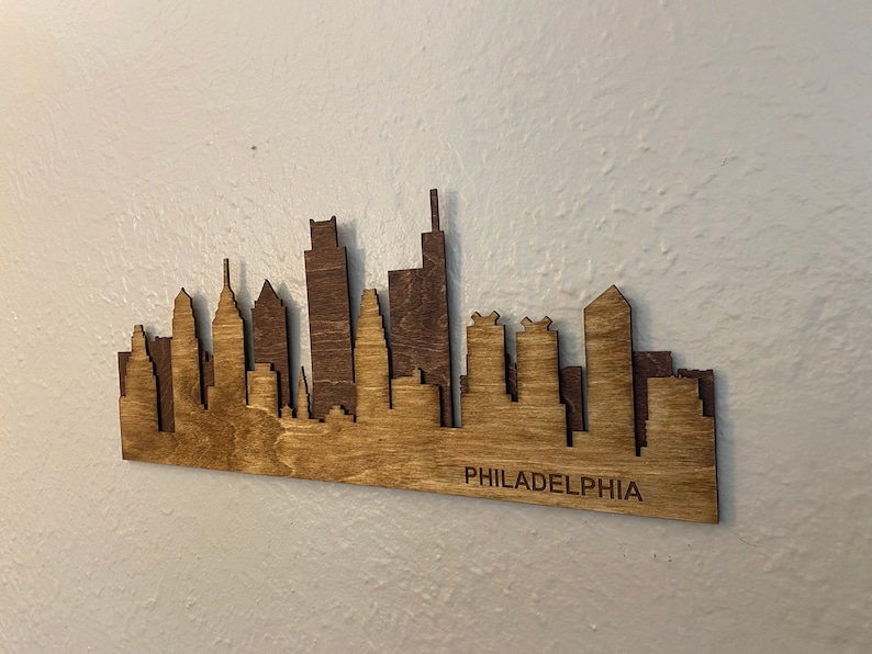 Puede incluir: Pieza de arte de madera con la silueta de Filadelfia. La silueta est&aacute; cortada con l&aacute;ser de madera, con un dise&ntilde;o de dos tonos, utilizando un marr&oacute;n m&aacute;s oscuro para algunos edificios. La palabra "PHILADELPHIA" est&aacute; grabada en la parte inferior.