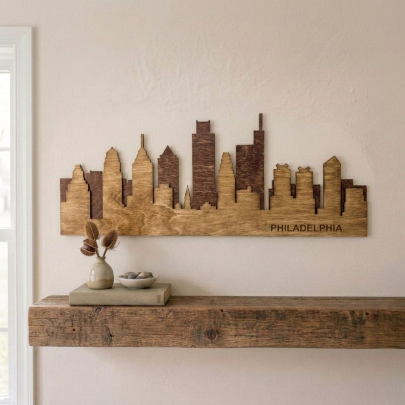 Puede incluir: Arte de pared de madera con la silueta de Filadelfia. El paisaje urbano est&aacute; cortado de madera, con un dise&ntilde;o de dos tonos: una base de madera m&aacute;s clara y edificios marrones m&aacute;s oscuros. La palabra "PHILADELPHIA" est&aacute; grabada en la base.