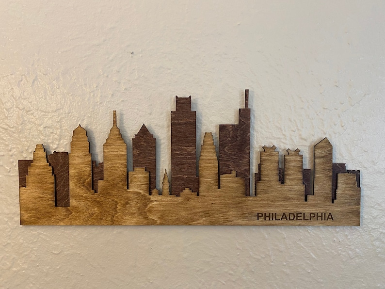 Puede incluir: Silueta de madera del horizonte de Filadelfia en dos tonos de marr&oacute;n. Los edificios de la ciudad, cortados con l&aacute;ser, est&aacute;n superpuestos, con la palabra "PHILADELPHIA" grabada en la parte inferior. El horizonte est&aacute; montado sobre una superficie clara.