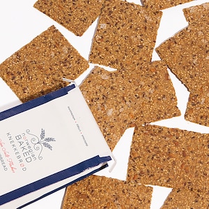 May include: A white and navy blue package of Norwegian Baked Knekkebr&oslash;d Sea Salt Flakes crispbread with several rectangular, seed-covered brown crackers scattered around it. The packaging features text including the product name and brand.