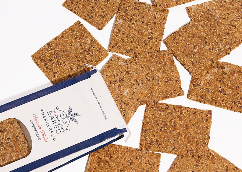 May include: A white and navy blue package of Norwegian Baked Knekkebr&oslash;d Sea Salt Flakes crispbread with several rectangular, seed-covered brown crackers scattered around it. The packaging features text including the product name and brand.