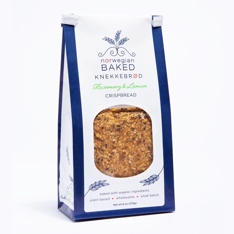 May include: A white and dark blue paper bag of Norwegian Baked Knekkebr&oslash;d Rosemary & Lemon crispbread. The front features a circular window revealing the textured, seeded crispbread inside. Text includes brand name, flavor, and net weight 6 oz (170g).