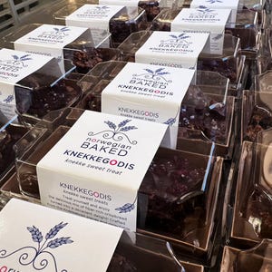 May include: Multiple clear plastic containers filled with dark brown, textured sweet treats. Each package features a white paper label with the text Norwegian Baked KNEKKEGODIS, a wheat stalk graphic, and additional descriptive product text.