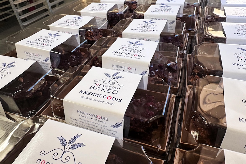 May include: Multiple clear plastic containers filled with dark brown, textured sweet treats. Each package features a white paper label with the text Norwegian Baked KNEKKEGODIS, a wheat stalk graphic, and additional descriptive product text.