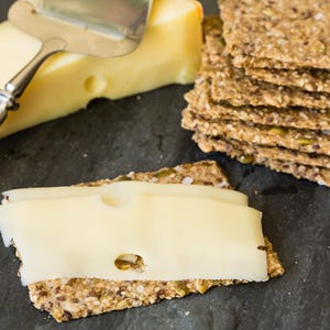 May include: A block of pale yellow Swiss cheese with visible holes, a stainless steel cheese slicer, and a stack of rectangular multi-grain crackers. One cracker is topped with a thin slice of the Swiss cheese, showing its texture and holes.