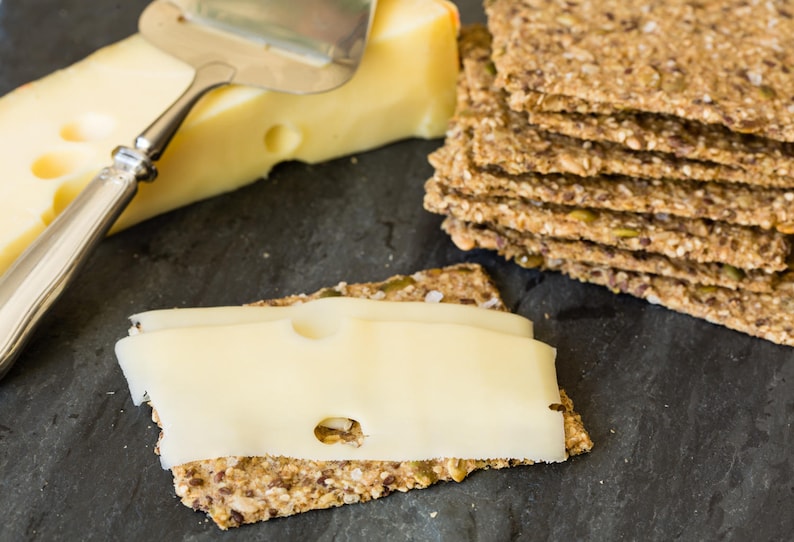 May include: A block of pale yellow Swiss cheese with visible holes, a stainless steel cheese slicer, and a stack of rectangular multi-grain crackers. One cracker is topped with a thin slice of the Swiss cheese, showing its texture and holes.
