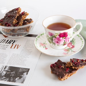 May include: A clear glass bowl filled with dark chocolate bark topped with dried cranberries, a floral teacup with tea, and two additional pieces of chocolate bark on a surface next to an open magazine featuring the text LEGENDARY LIV.