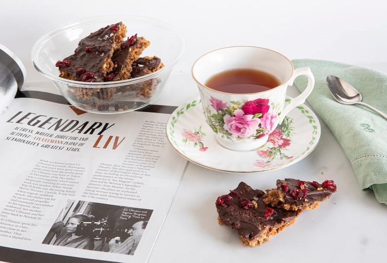 May include: A clear glass bowl filled with dark chocolate bark topped with dried cranberries, a floral teacup with tea, and two additional pieces of chocolate bark on a surface next to an open magazine featuring the text LEGENDARY LIV.