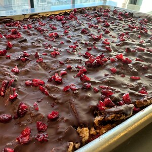 May include: A rectangular metal baking sheet filled with a crispy treat base topped with a thick layer of smooth dark chocolate and scattered pieces of bright red dried cranberries, creating a textured surface across the entire pan.