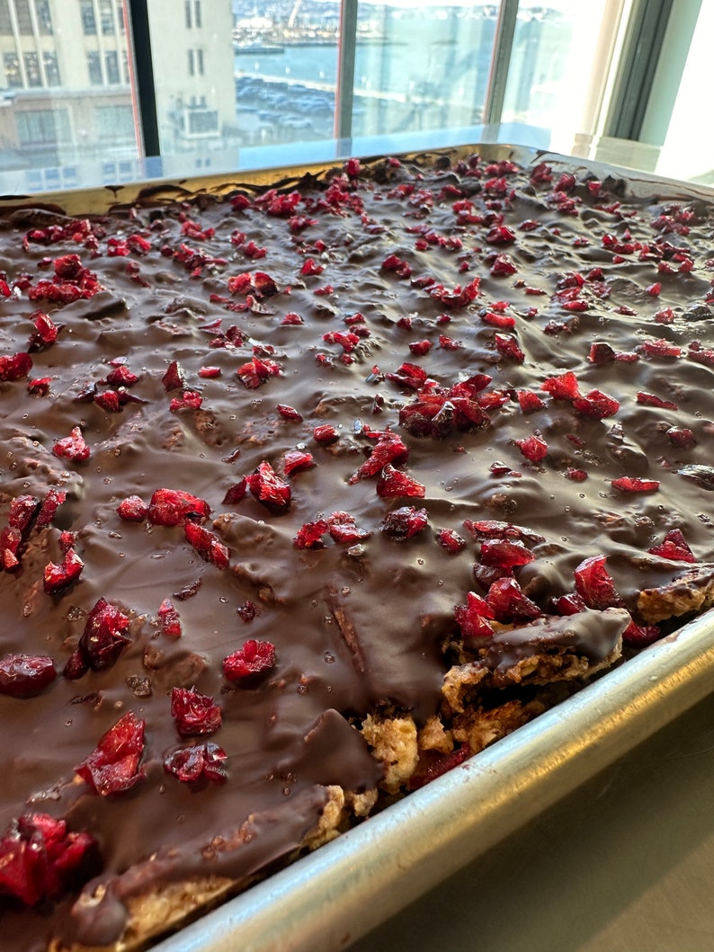 May include: A rectangular metal baking sheet filled with a crispy treat base topped with a thick layer of smooth dark chocolate and scattered pieces of bright red dried cranberries, creating a textured surface across the entire pan.