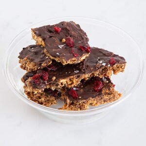 May include: A clear glass bowl filled with several broken pieces of dark chocolate-covered granola bark. Each piece features a textured oat base topped with a smooth layer of dark chocolate, scattered dried red cranberries, and coarse sea salt.