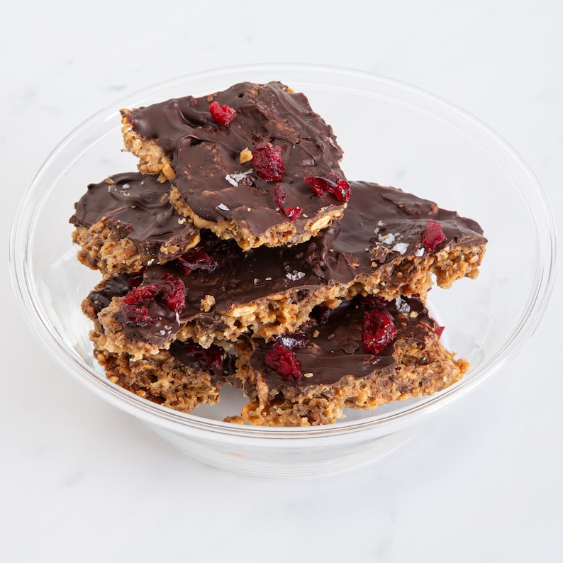 May include: A clear glass bowl filled with several broken pieces of dark chocolate-covered granola bark. Each piece features a textured oat base topped with a smooth layer of dark chocolate, scattered dried red cranberries, and coarse sea salt.