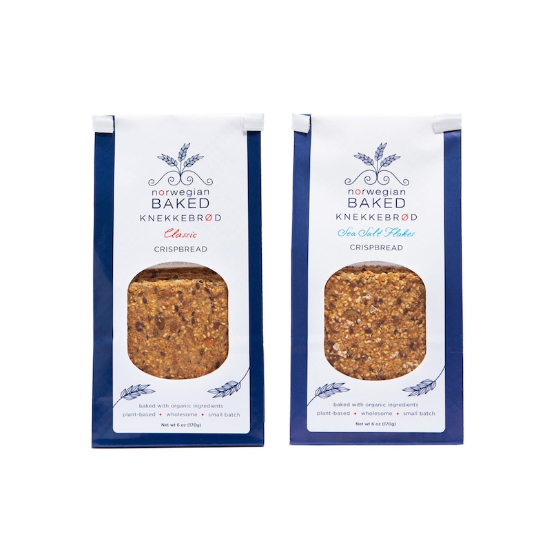 May include: Two white and blue vertical bags of Norwegian Baked Knekkebr&oslash;d crispbread. The left bag is labeled Classic and the right bag is labeled Sea Salt Flakes. Each bag features a clear window showing the textured, seeded crispbread inside.