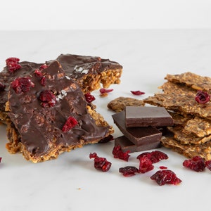 May include: A collection of oat-based brittle pieces topped with dark chocolate and dried cranberries, accompanied by three stacked dark chocolate squares and scattered dried cranberries on a smooth white surface.