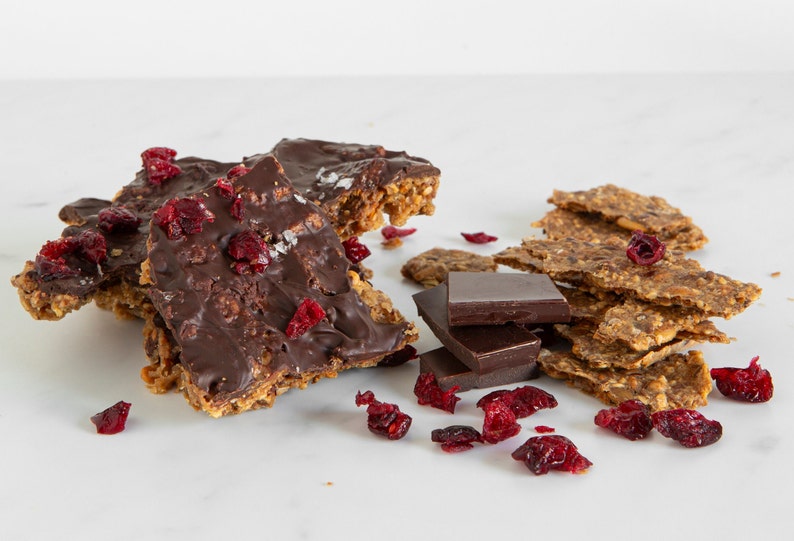 May include: A collection of oat-based brittle pieces topped with dark chocolate and dried cranberries, accompanied by three stacked dark chocolate squares and scattered dried cranberries on a smooth white surface.