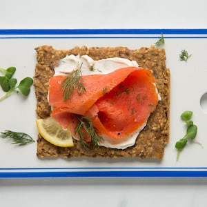 May include: A rectangular whole grain crispbread topped with white cream cheese, bright orange smoked salmon, a thin slice of yellow lemon, and fresh green dill sprigs, presented on a white rectangular serving board with a blue border.