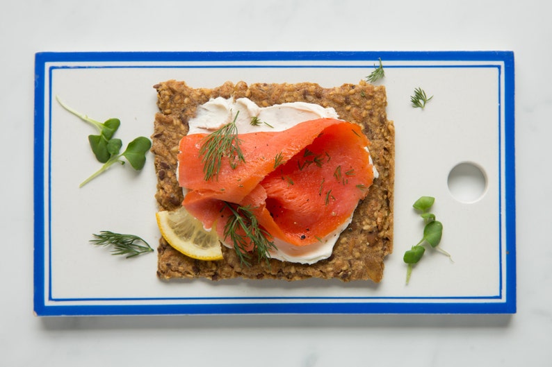 May include: A rectangular whole grain crispbread topped with white cream cheese, bright orange smoked salmon, a thin slice of yellow lemon, and fresh green dill sprigs, presented on a white rectangular serving board with a blue border.
